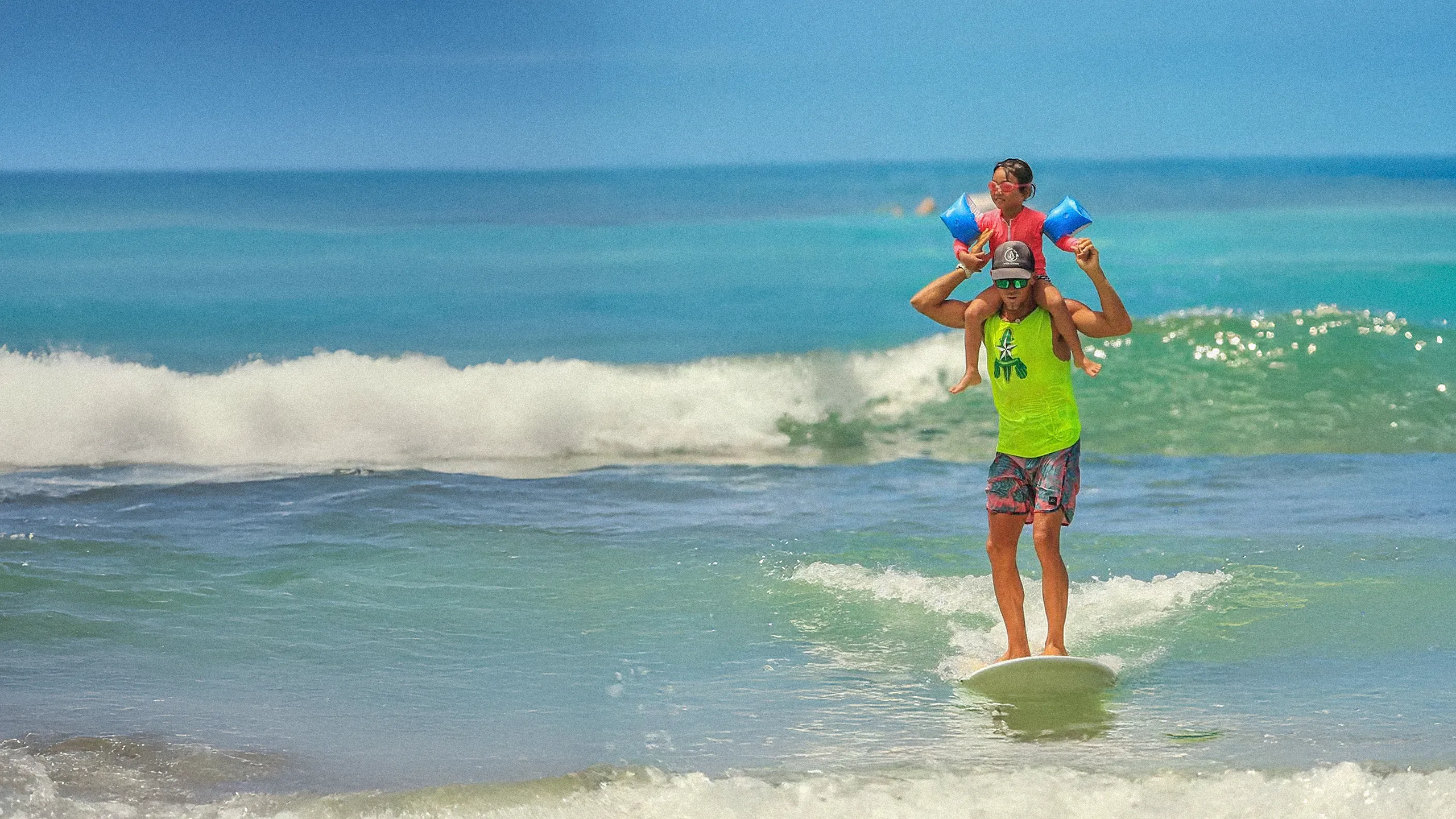 Man in neon green shirt surfing with child wearing floaties on his shoulders in ocean waves under clear blue sky.