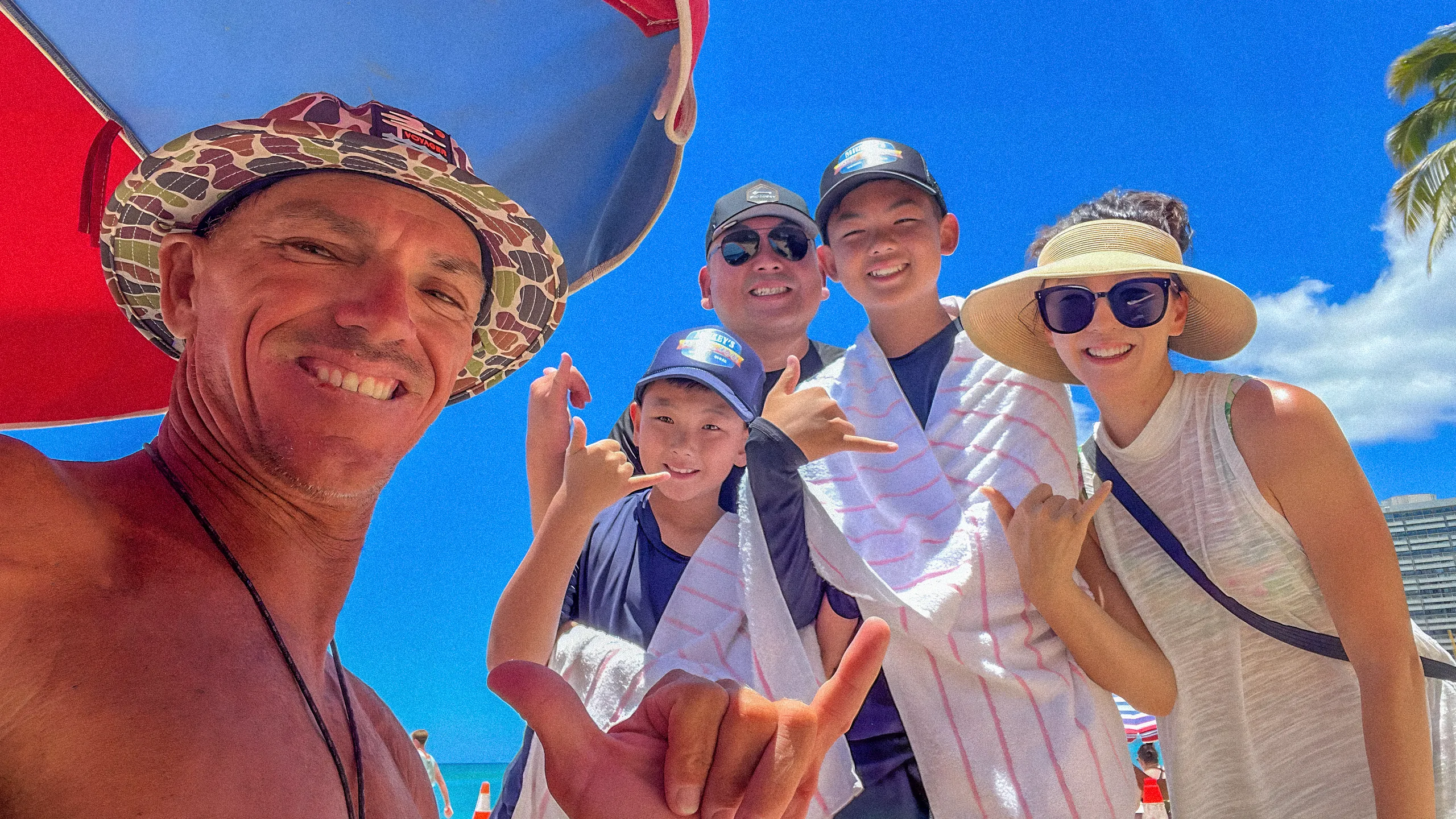 Group of five smiling people at the beach under a red umbrella, wearing hats and sunglasses, making shaka hand signs with blue sky and palm tree in the background.