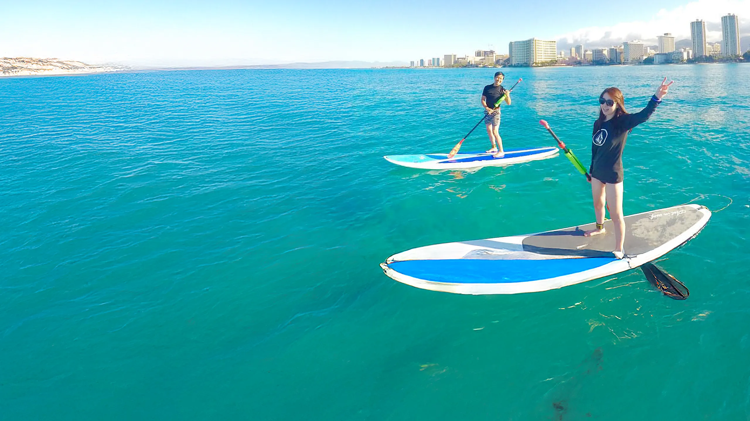 Two people paddleboarding on clear blue ocean water near a city skyline on a sunny day.