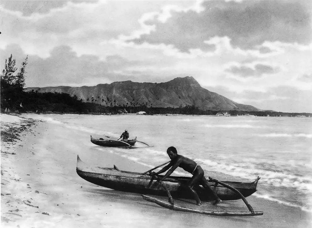 Historic photograph of two Indigenous Hawaiians with outrigger canoes on a shoreline in Honolulu, Hawaii.